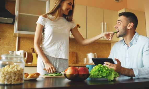 Attractive couple chatting in the kitchen early morning. Beautiful woman feed her husband while cooking breakfast at home